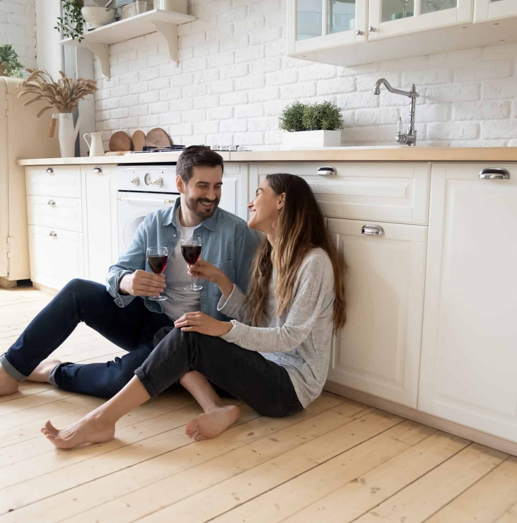 Romantic couple sit on warm kitchen floor talking drinking wine - behind them is a custom wooden counter top and custom white cabinetry
